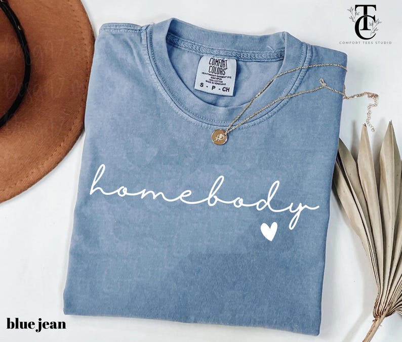 May include: A blue jean t-shirt with the text "homebody" in white script with a small white heart underneath. The shirt has a distressed look and is folded in half.