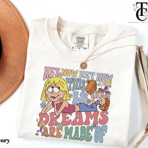 May include: Ivory-colored t-shirt with a cartoon graphic. The graphic features a blonde character with the text "HEY NOW HEY NOW THIS IS WHAT DREAMS ARE MADE OF" in colorful bubble letters. A gold necklace is draped across the shirt.