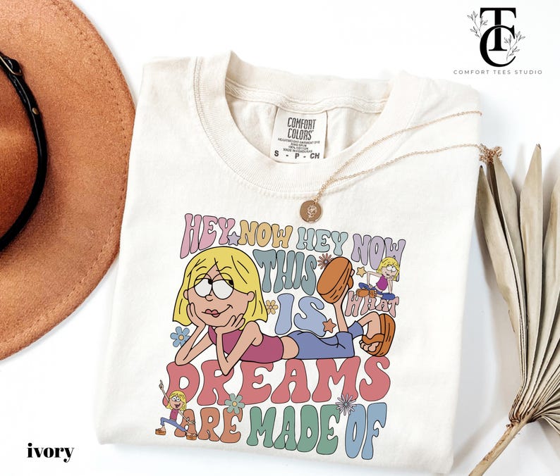 May include: Ivory-colored t-shirt with a cartoon graphic. The graphic features a blonde character with the text "HEY NOW HEY NOW THIS IS WHAT DREAMS ARE MADE OF" in colorful bubble letters. A gold necklace is draped across the shirt.