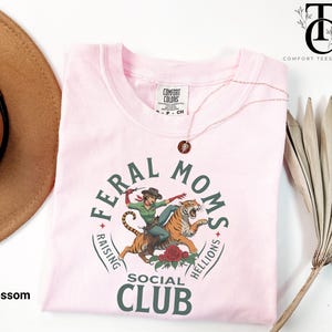 May include: Pink t-shirt with a graphic of a woman riding a tiger. The graphic is surrounded by a circle with the text "Feral Moms Raising Hellions Social Club".