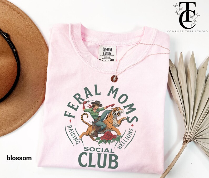 May include: Pink t-shirt with a graphic of a woman riding a tiger. The graphic is surrounded by a circle with the text "Feral Moms Raising Hellions Social Club".