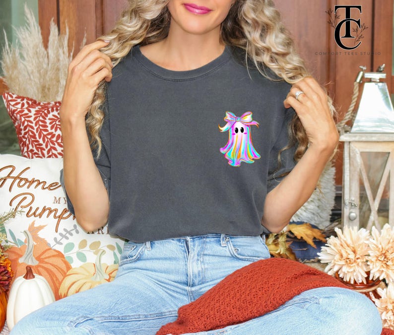 May include: Dark gray t-shirt featuring a colorful ghost design with a rainbow bow. The ghost has a friendly expression. The shirt is a comfortable, casual style, perfect for Halloween or everyday wear. The Comfort Tee Studio logo is visible in the upper right corner.