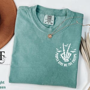 May include: Light green t-shirt with the text "People Give Me The Creeps" in white, featuring a skeleton hand making a peace sign with bats. A gold necklace with a pendant is on the shirt.