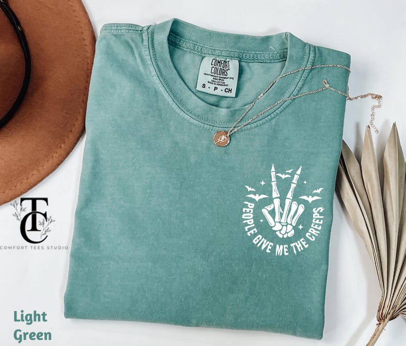 May include: Light green t-shirt with the text "People Give Me The Creeps" in white, featuring a skeleton hand making a peace sign with bats. A gold necklace with a pendant is on the shirt.