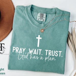 May include: Light green t-shirt with a white cross and the text "PRAY. WAIT. TRUST. God has a plan".