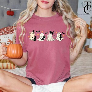 Op de afbeelding: Een bordeauxrood T-shirt met een Halloween-ontwerp met vier cartoongeesten, zwarte katten, vleermuizen en sterren. Het shirt heeft een comfortabele pasvorm en is geschikt voor het herfstseizoen. Een persoon houdt een oranje pompoenvormige mok vast.