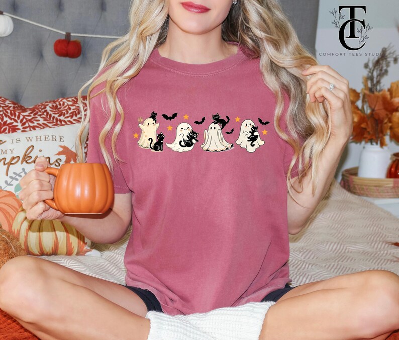 Op de afbeelding: Een bordeauxrood T-shirt met een Halloween-ontwerp met vier cartoongeesten, zwarte katten, vleermuizen en sterren. Het shirt heeft een comfortabele pasvorm en is geschikt voor het herfstseizoen. Een persoon houdt een oranje pompoenvormige mok vast.