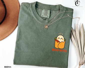 May include: Moss green t-shirt with a "Boo-Rito" graphic. The design features a cartoon ghost inside a burrito, with orange text below. A gold necklace and a brown hat are also visible.