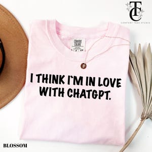 May include: A light pink t-shirt with the text "I THINK I'M IN LOVE WITH CHATGPT." in black. The shirt has a crew neck and a Comfort Colors tag. A delicate necklace with a pendant rests on the shirt.