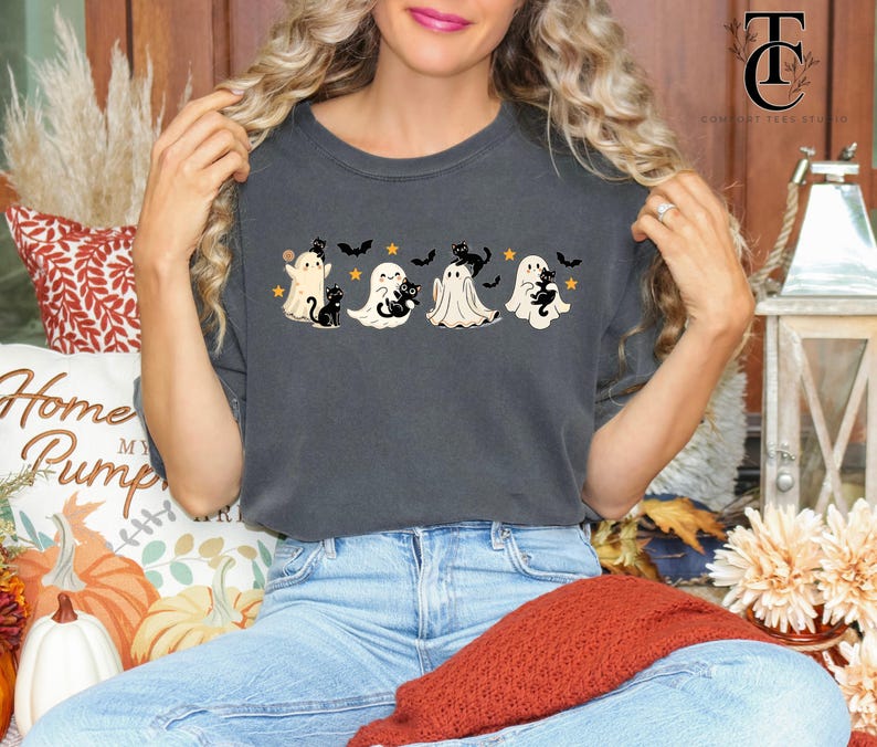 Op de afbeelding: Donkergrijs T-shirt met een Halloween-ontwerp. Het ontwerp bevat vier geesten, zwarte katten, vleermuizen en sterren. De afbeelding is over de borst gedrukt. Een comfortabele, casual stijl.