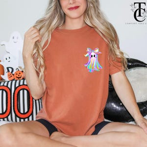 May include: Rust-colored t-shirt featuring a colorful ghost graphic with a bow. The ghost design has rainbow hues. The shirt is part of a Halloween-themed collection. The Comfort Tees Studio logo is visible in the upper right corner.