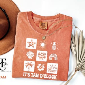 May include: A coral orange t-shirt with a white graphic design of summer icons and the text "It's Tan O'Clock".