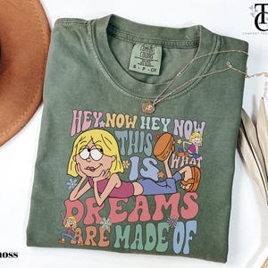 May include: Moss green t-shirt with a graphic of a cartoon character and the text "HEY NOW HEY NOW THIS IS WHAT DREAMS ARE MADE OF". The shirt has a crew neck and short sleeves. A gold necklace is draped across the shirt.
