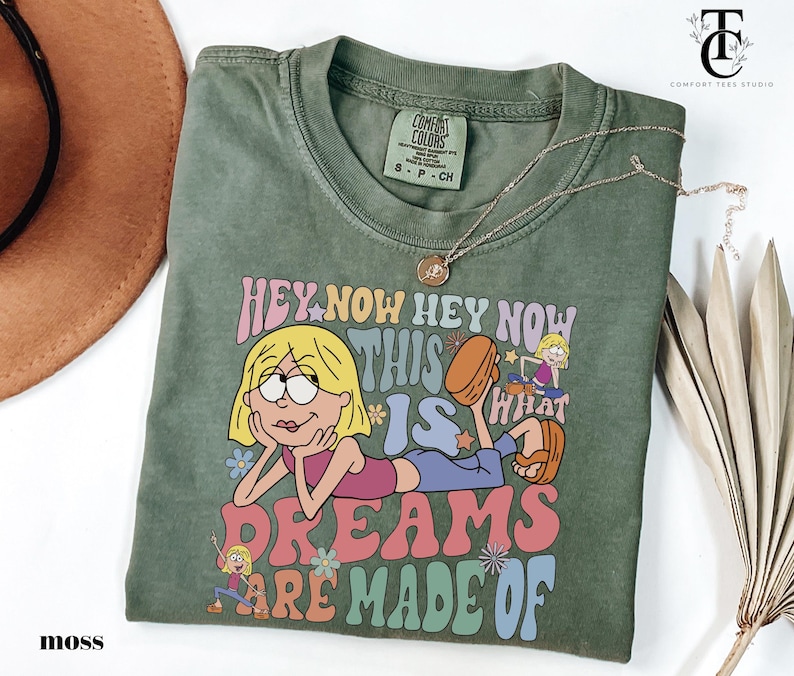 May include: Moss green t-shirt with a graphic of a cartoon character and the text "HEY NOW HEY NOW THIS IS WHAT DREAMS ARE MADE OF". The shirt has a crew neck and short sleeves. A gold necklace is draped across the shirt.