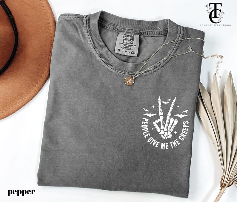 May include: A gray t-shirt with a crew neck and short sleeves. The shirt features a white graphic of a skeleton hand making a peace sign, with the text "PEOPLE GIVE ME THE CREEPS". A silver necklace with a pendant is on the shirt.