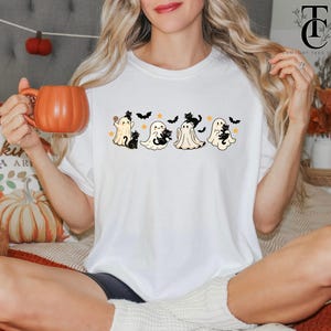 Op de afbeelding: Wit T-shirt met een Halloween-ontwerp met cartoongeesten, zwarte katten, sterren en vleermuizen. Het ontwerp is gecentreerd op het shirt. De persoon houdt een oranje pompoenvormige mok vast.