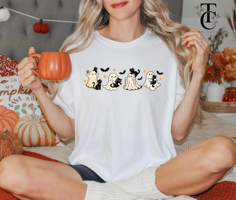 Op de afbeelding: Wit T-shirt met een Halloween-ontwerp met cartoongeesten, zwarte katten, sterren en vleermuizen. Het ontwerp is gecentreerd op het shirt. De persoon houdt een oranje pompoenvormige mok vast.