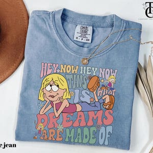 May include: A light blue t-shirt with the text "HEY NOW HEY NOW THIS IS WHAT DREAMS ARE MADE OF" and a cartoon illustration. The shirt has a crew neck and short sleeves. A gold necklace with a pendant is draped across the shirt.