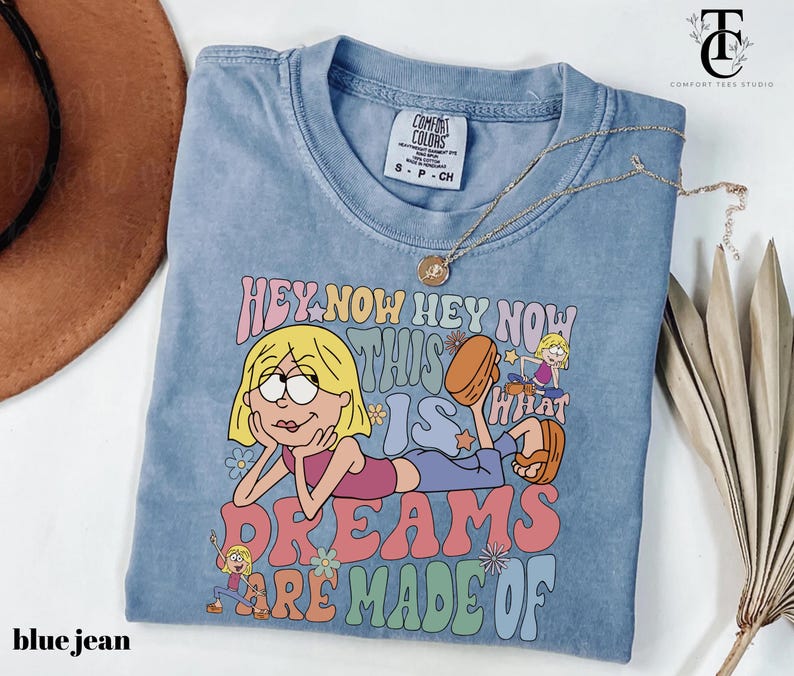 May include: A light blue t-shirt with the text "HEY NOW HEY NOW THIS IS WHAT DREAMS ARE MADE OF" and a cartoon illustration. The shirt has a crew neck and short sleeves. A gold necklace with a pendant is draped across the shirt.