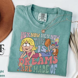 May include: Light green t-shirt with the text "HEY NOW HEY NOW THIS IS WHAT DREAMS ARE MADE OF" and a cartoon graphic. The shirt has a crew neck and short sleeves. A gold necklace is draped across the shirt.