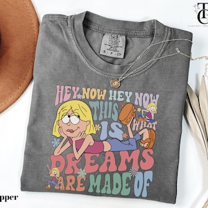 May include: A gray t-shirt with a graphic featuring a cartoon character and the text "HEY NOW HEY NOW THIS IS WHAT DREAMS ARE MADE OF." The shirt has a crew neck and short sleeves. A silver necklace with a pendant is draped across the shirt.