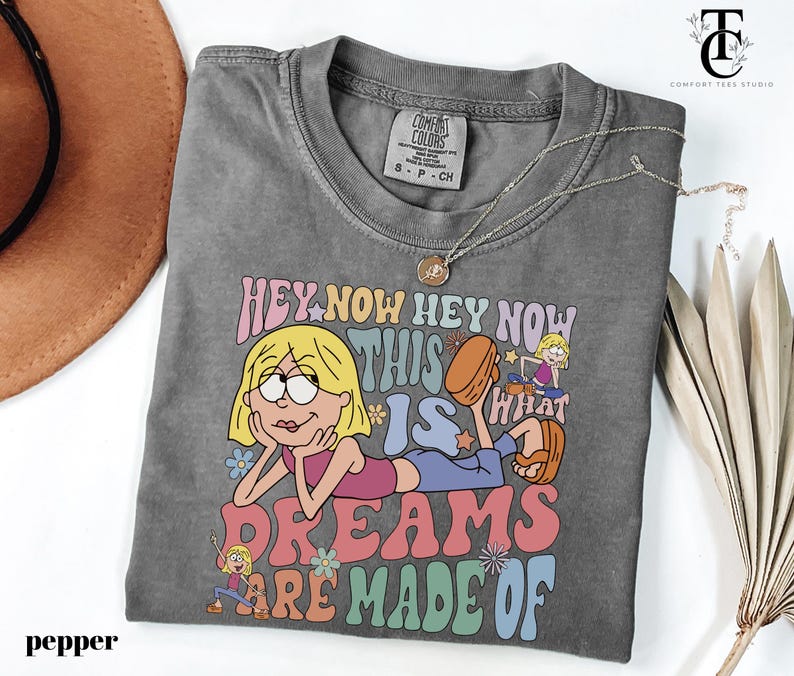 May include: A gray t-shirt with a graphic featuring a cartoon character and the text "HEY NOW HEY NOW THIS IS WHAT DREAMS ARE MADE OF." The shirt has a crew neck and short sleeves. A silver necklace with a pendant is draped across the shirt.
