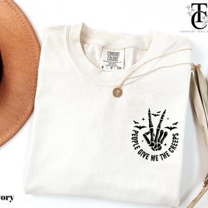 May include: Ivory-colored t-shirt with a black graphic of a skeleton hand making a peace sign, surrounded by bats and the text "PEOPLE GIVE ME THE CREEPS." A gold necklace and a brown hat are also visible.