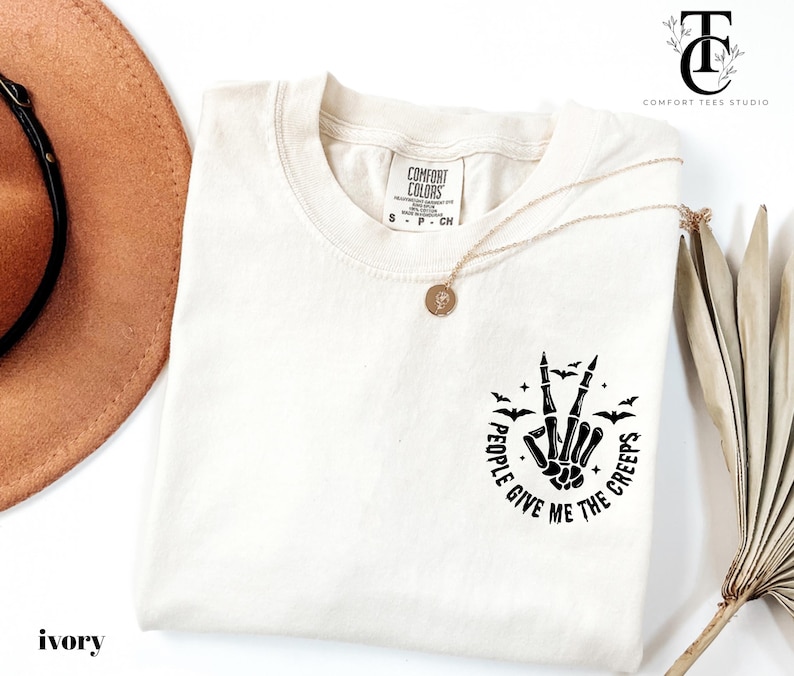 May include: Ivory-colored t-shirt with a black graphic of a skeleton hand making a peace sign, surrounded by bats and the text "PEOPLE GIVE ME THE CREEPS." A gold necklace and a brown hat are also visible.