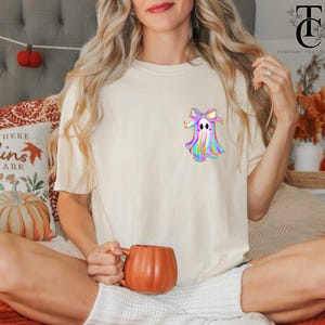 May include: Cream-colored t-shirt featuring a colorful ghost graphic with a bow. The ghost design has a rainbow color scheme. The person is holding an orange pumpkin-shaped mug. The shirt is part of a fall-themed collection.