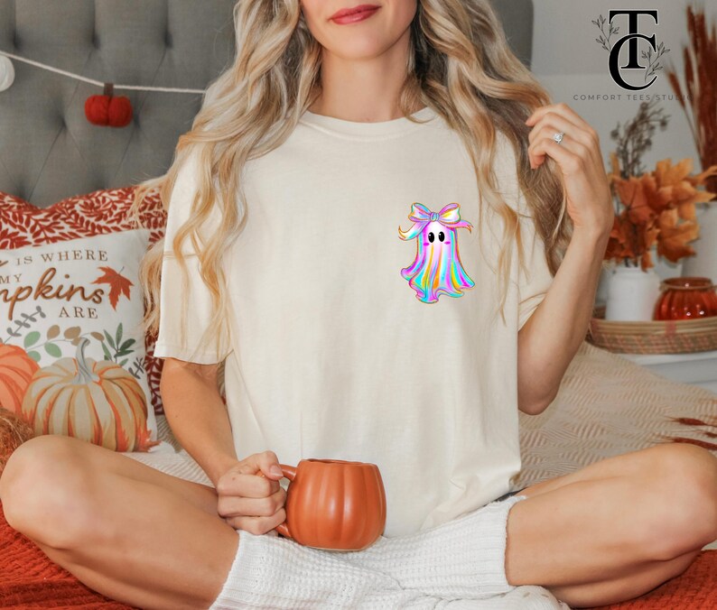 May include: Cream-colored t-shirt featuring a colorful ghost graphic with a bow. The ghost design has a rainbow color scheme. The person is holding an orange pumpkin-shaped mug. The shirt is part of a fall-themed collection.