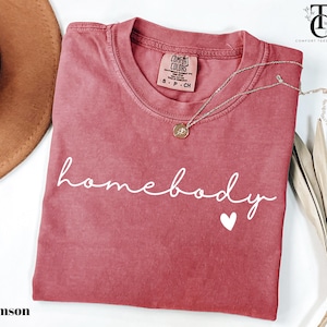 May include: Crimson red t-shirt with the text "homebody" in white script font and a small white heart. The shirt is made of a soft, comfortable material.