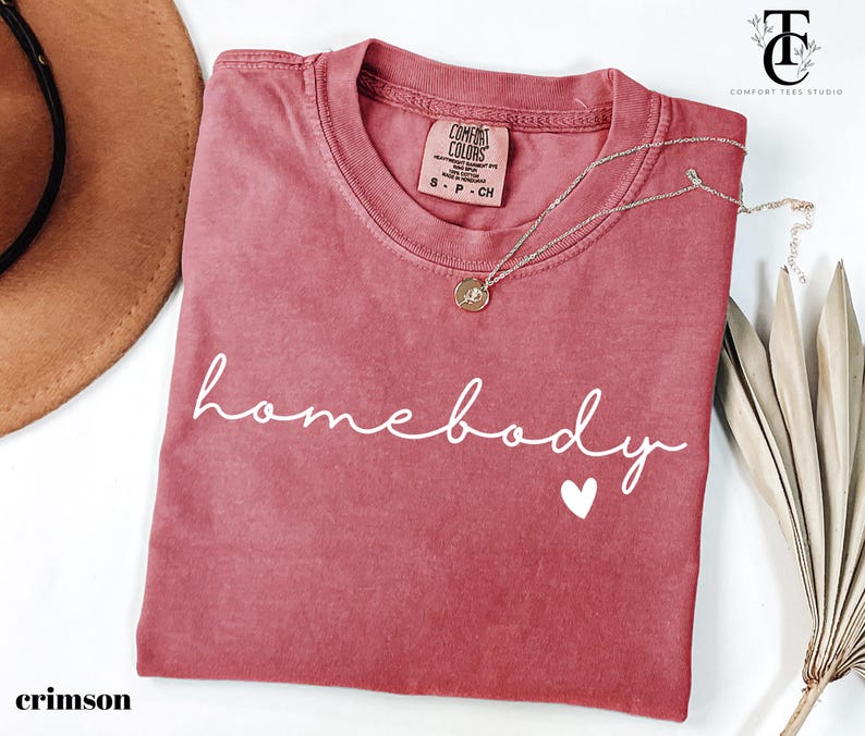 May include: Crimson red t-shirt with the text "homebody" in white script font and a small white heart. The shirt is made of a soft, comfortable material.