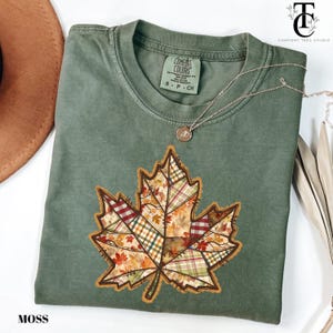 May include: A moss green t-shirt featuring a large, patchwork-style maple leaf design. The leaf incorporates various patterns and colors, including plaid and autumn hues. A silver necklace with a pendant is draped across the shirt. The word "MOSS" is printed at the bottom.