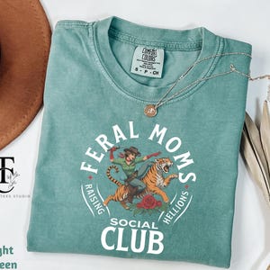 May include: Light green t-shirt with a graphic of a woman riding a tiger. The graphic includes the text "Feral Moms Raising Hellions Social Club".