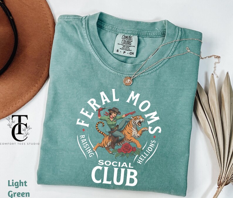 May include: Light green t-shirt with a graphic of a woman riding a tiger. The graphic includes the text "Feral Moms Raising Hellions Social Club".