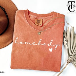 May include: A coral-coloured t-shirt with the text "homebody" in white script with a small white heart below. The shirt is made of a soft, comfortable material and has a relaxed fit.