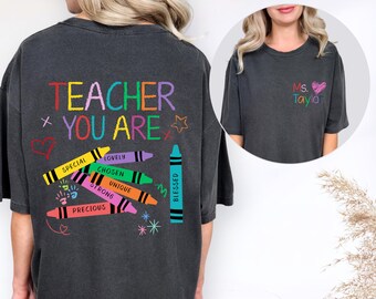 May include: A charcoal gray t-shirt featuring a back graphic with the text TEACHER YOU ARE in colorful letters, accompanied by illustrations of crayons labeled SPECIAL, LOVELY, CHOSEN, UNIQUE, STRONG, PRECIOUS, and BLESSED. The front chest displays Ms. Taylor.