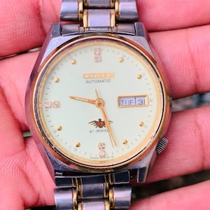 May include: A Citizen automatic wristwatch with a silver and gold-tone metal band. The watch face is cream-colored with gold hands and hour markers. The date is displayed on the dial. The watch has a classic, vintage aesthetic.
