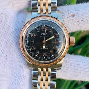 Puede incluir: Un reloj de pulsera con esfera negra, agujas doradas y visualización de la fecha. El reloj tiene una correa de acero inoxidable y dorada. La esfera tiene el texto "ORIS 25 JEWELS AUTOMATIC SWISS MADE".