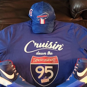 May include: Blue t-shirt with the text "Cruisin' down the Interstate 95" printed on the front. The shirt is paired with a blue baseball cap with a Buffalo Bills logo and a pair of blue and white sneakers with red accents.