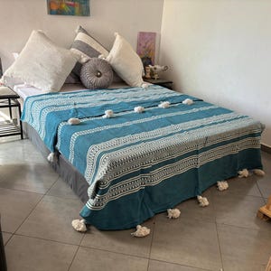 Puede incluir: Una manta de tiro a rayas azul turquesa y blanca con pompones blancos en los bordes. La manta está colocada sobre una cama con almohadas blancas.