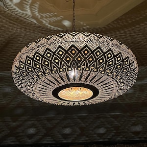 May include: A large, circular, dark-colored metal pendant light fixture featuring intricate, laser-cut geometric patterns. The design includes a central opening and a series of radiating, diamond-shaped cutouts that cast detailed light patterns.
