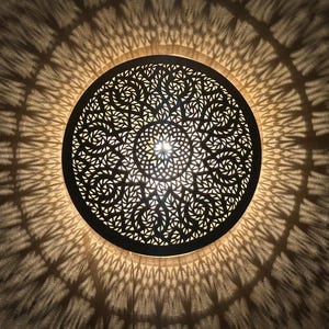 May include: A round, black metal ceiling light fixture with an intricate, floral-like cutout design. The light casts a decorative shadow pattern on the surrounding surface, creating a warm, inviting ambiance. The fixture is centered in the image.