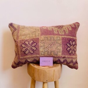 May include: A rectangular pillow with a geometric pattern in shades of beige, pink, and purple. The pillow is made of woven fabric and has a fringed edge. The pillow is sitting on a wooden stool with a purple tag that says "CRAFT IT" on it.
