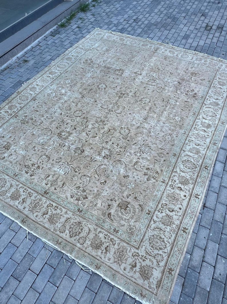 K&ouml;nnte beinhalten: Ein Vintage-Teppich mit beigefarbenem Hintergrund und einem floralen Muster in Braun- und Gr&uuml;nt&ouml;nen. Der Teppich hat einen Rand mit einem sich wiederholenden Muster.