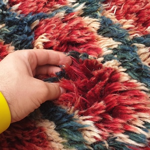 May include: A hand touches a colorful rug with a pattern of red, blue, and cream. The rug has a thick, textured pile. A yellow bracelet is on the person's wrist.