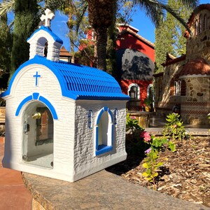 Puede incluir: Una peque&ntilde;a capilla blanca y azul con una cruz en la parte delantera y una campana en el interior. La capilla est&aacute; hecha de piedra y tiene un techo azul. Est&aacute; sentada en un muro de piedra frente a una fuente.