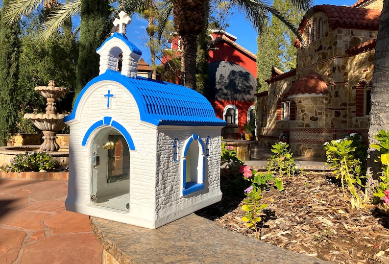 Puede incluir: Una peque&ntilde;a capilla blanca y azul con una cruz en la parte delantera y una campana en el interior. La capilla est&aacute; hecha de piedra y tiene un techo azul. Est&aacute; sentada en un muro de piedra frente a una fuente.