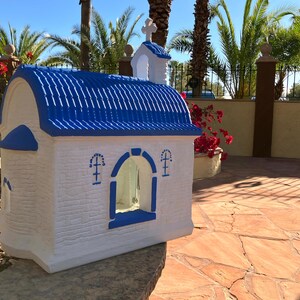 Puede incluir: Una peque&ntilde;a figura de iglesia blanca y azul con un techo azul y una ventana con un borde azul. La figura est&aacute; sentada sobre una superficie de piedra.
