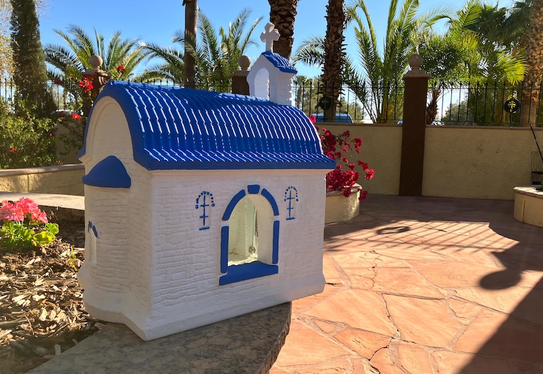 Puede incluir: Una peque&ntilde;a figura de iglesia blanca y azul con un techo azul y una ventana con un borde azul. La figura est&aacute; sentada sobre una superficie de piedra.